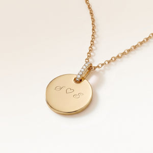 Close-up of a round gold pendant with a zirconia-set bail and personalised engraving of initials with a heart