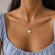 Model in a light blue top wearing a personalized heart-shaped silver necklace with white zircons and an engraved date, with space for engraving of text, coordinates or handwriting on the front and back