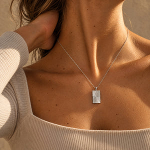 Model in an oatmeal rib-knit top wearing a square silver necklace with a ridged sunburst pattern and room for personal engraving of text, coordinates or handwriting on the back
